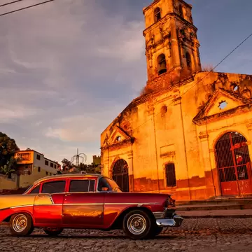 Ruined colonial church with a tall bell tower glowing at sunset, a classic car parked on cobbles.