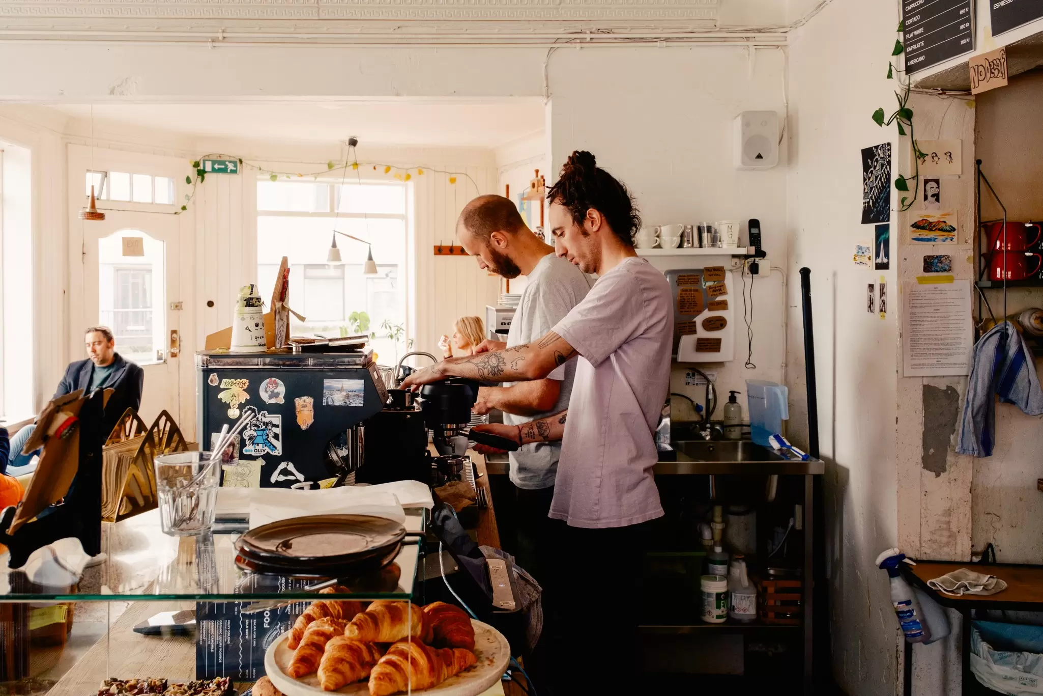 Two men prepare coffee drinks behind the counter of a cafe.