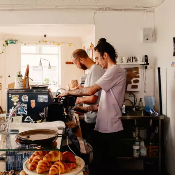 Two men prepare coffee drinks behind the counter of a cafe.