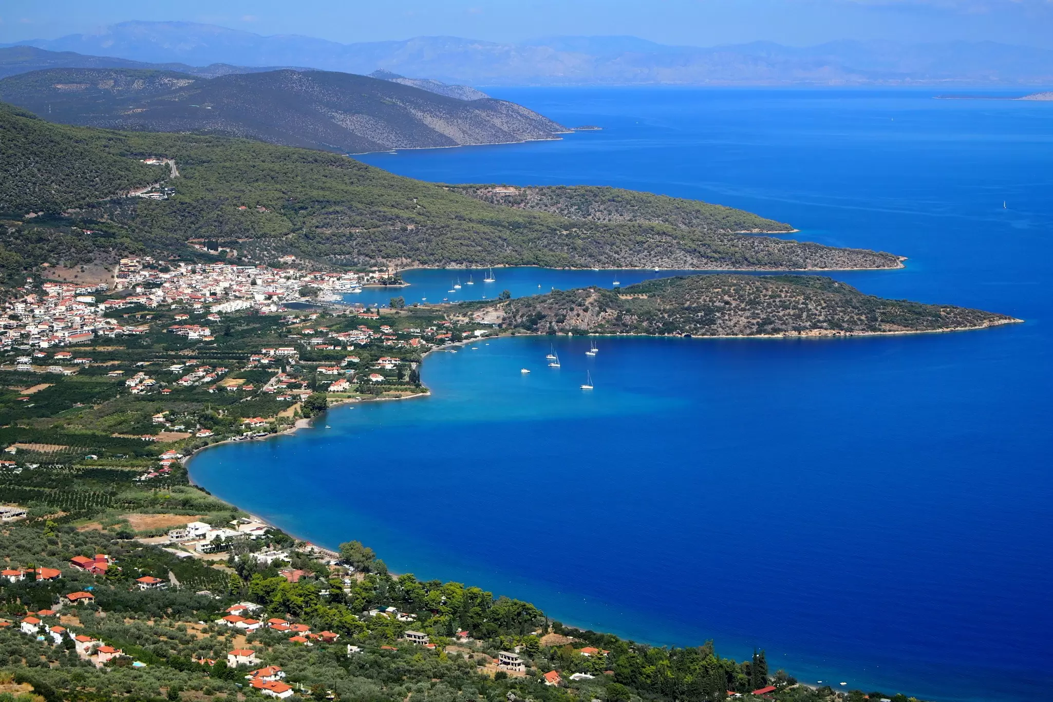 An aerial view of a gulf with small towns dotting the green coastline in Greece. Blue water and sunshine. 