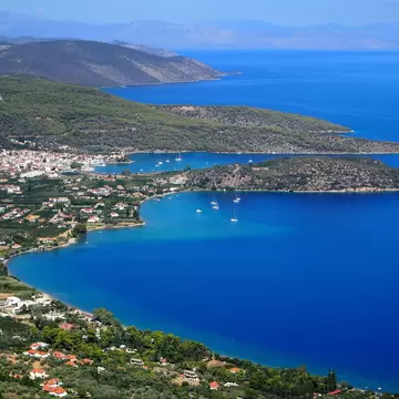 An aerial view of a gulf with small towns dotting the green coastline in Greece. Blue water and sunshine. 