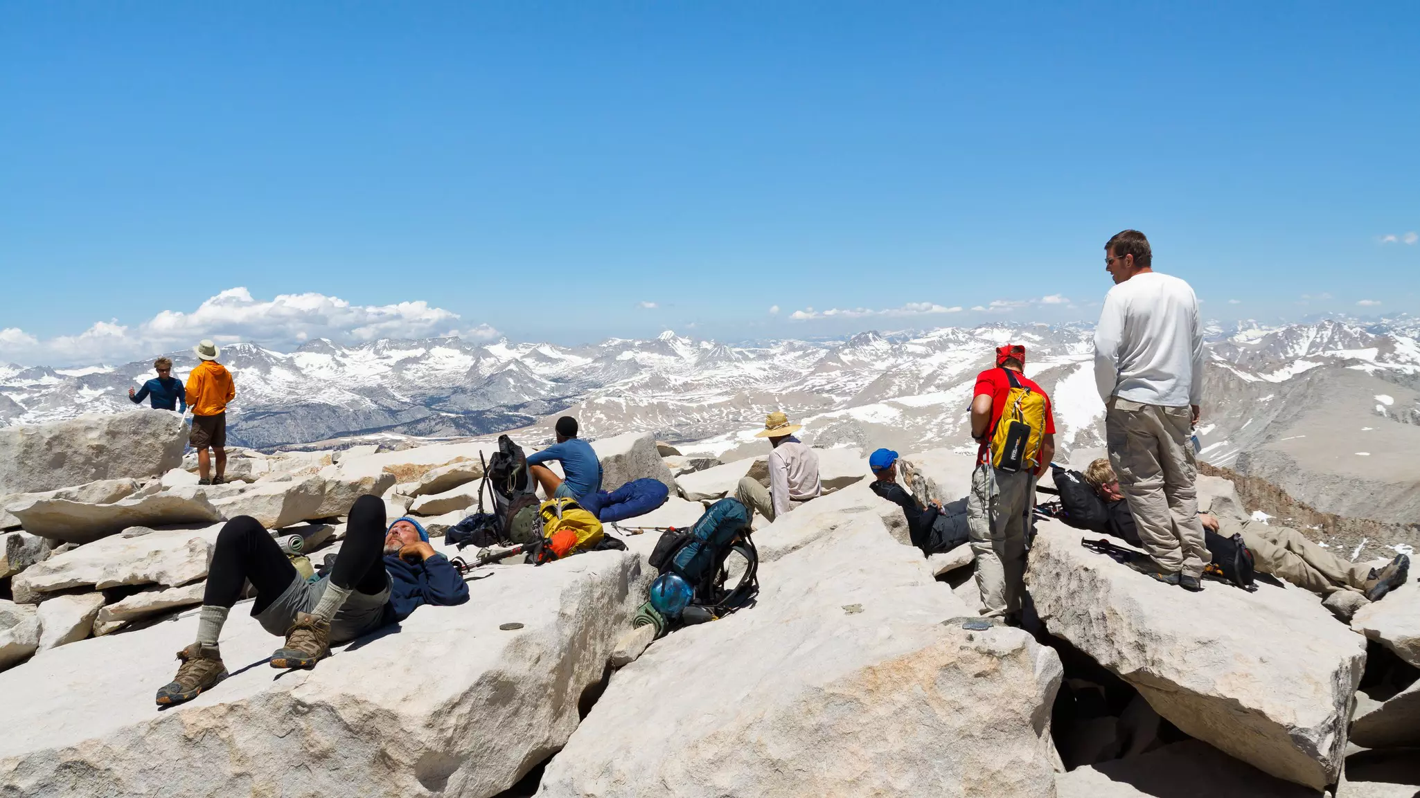 A group of hikers lie on rocks and rest at the top of a mountain, with views of snow-capped peaks in the distance.