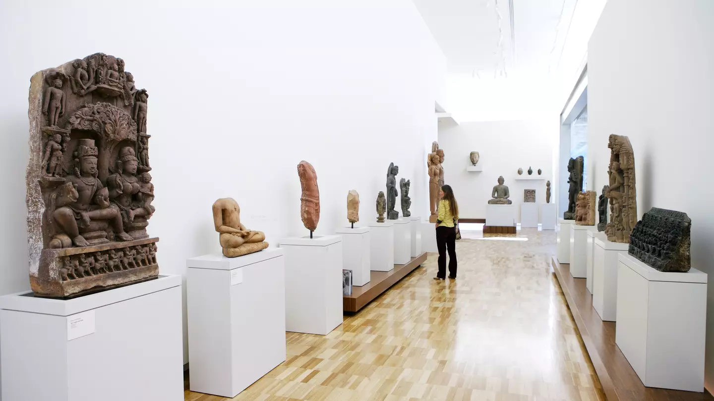 View of a long hallway with sculptures on either side. A woman is looking at a sculpture on one side of the hallway.