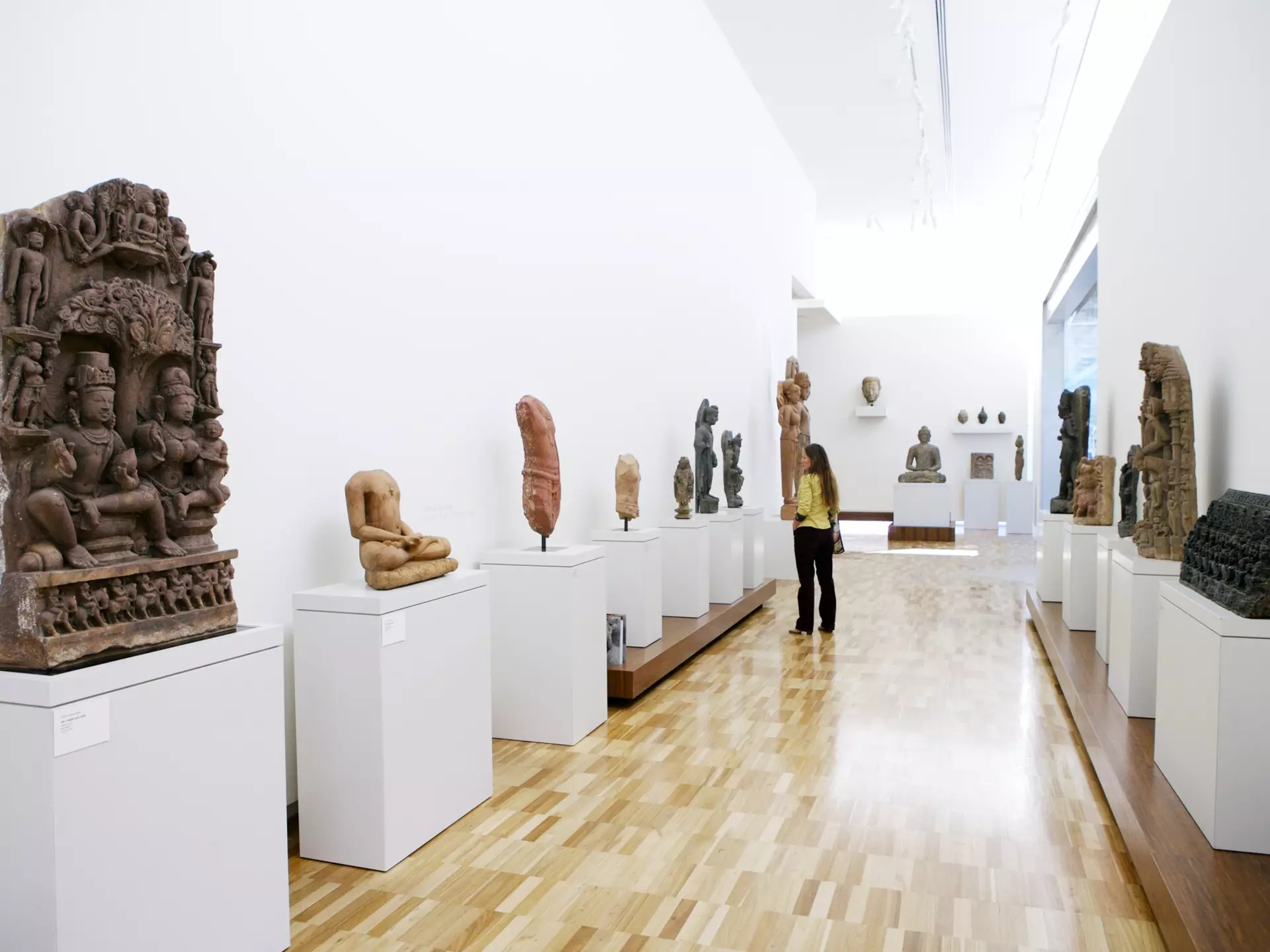 View of a long hallway with sculptures on either side. A woman is looking at a sculpture on one side of the hallway.