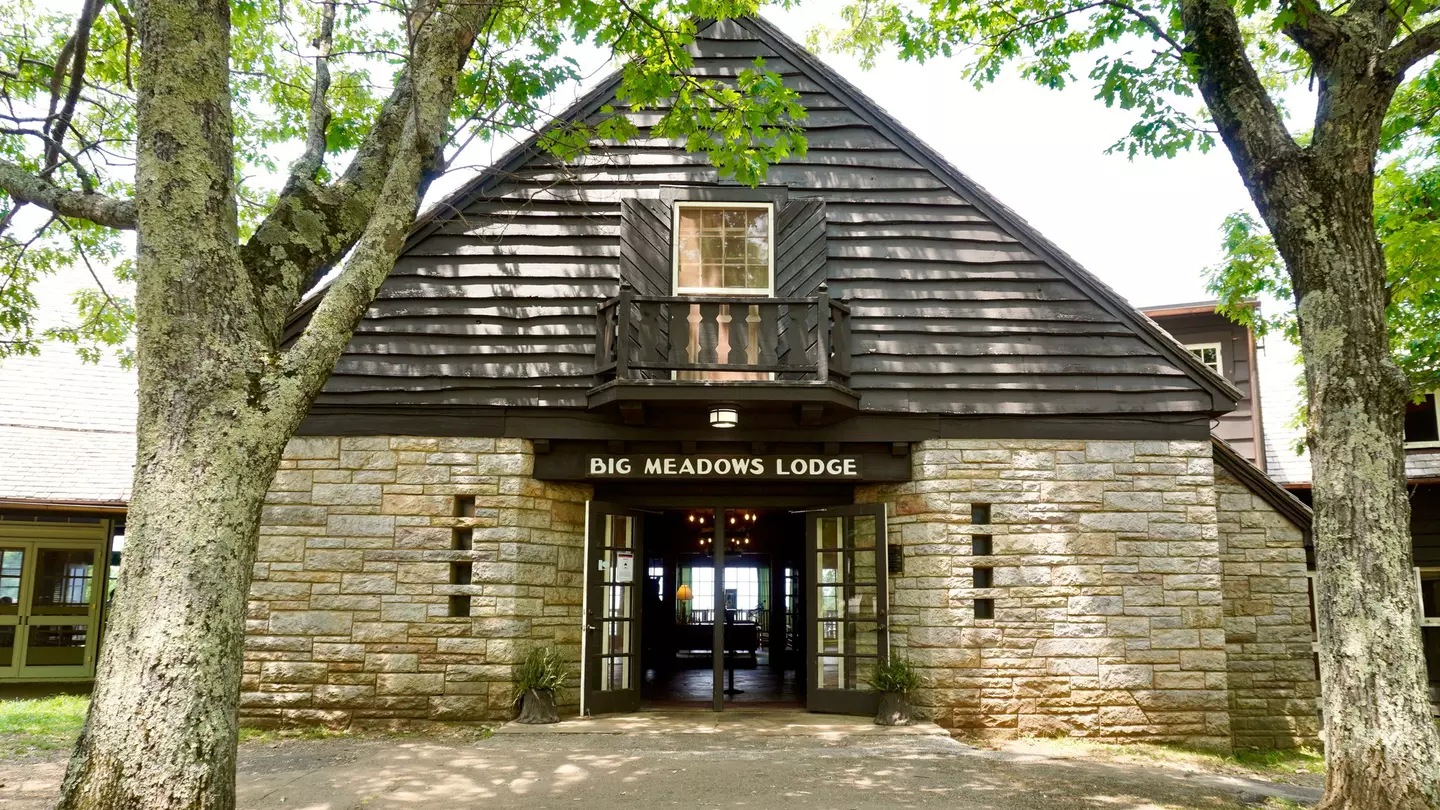 Photo of stone lodge with clapboard siding with a brown side above open doors reading "Big Meadows Lodge" in white capital letter. Sunlight casts shadows through deciduous trees onto a dirt walkway in the foreground.
