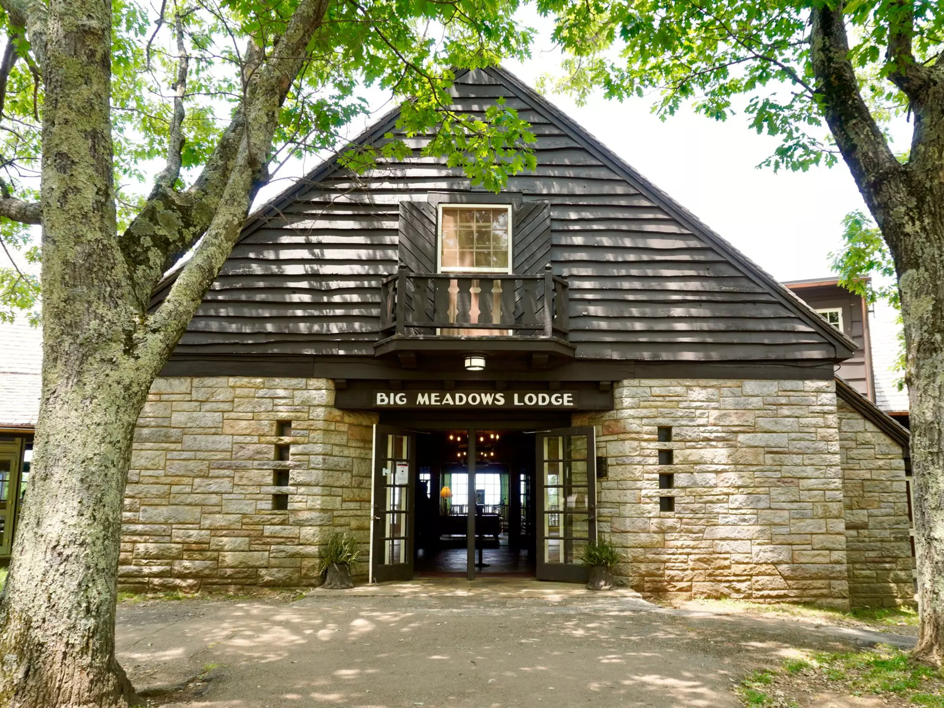 Photo of stone lodge with clapboard siding with a brown side above open doors reading "Big Meadows Lodge" in white capital letter. Sunlight casts shadows through deciduous trees onto a dirt walkway in the foreground.