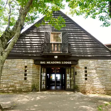 Photo of stone lodge with clapboard siding with a brown side above open doors reading "Big Meadows Lodge" in white capital letter. Sunlight casts shadows through deciduous trees onto a dirt walkway in the foreground.