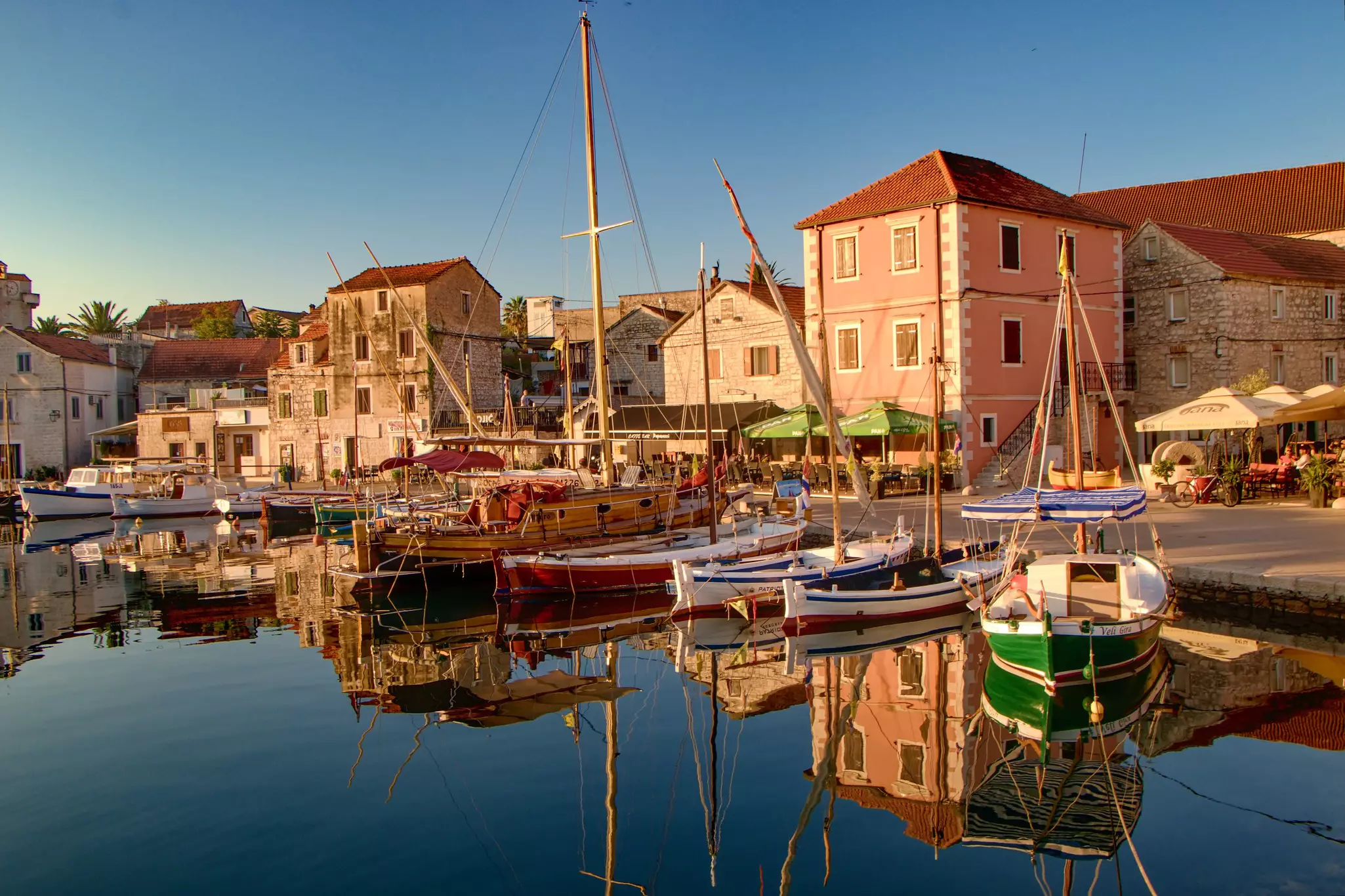Boats rest at a harbor during sunrise, with buildings at the port. The scene is reflected in the still water.