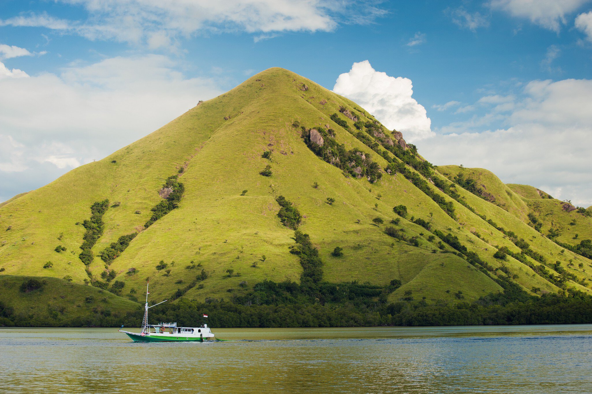 The sun strikes the green grassy slopes of Rinca Island (also known as Rincah and Rindja), as a small boat travels through the water in the foreground.