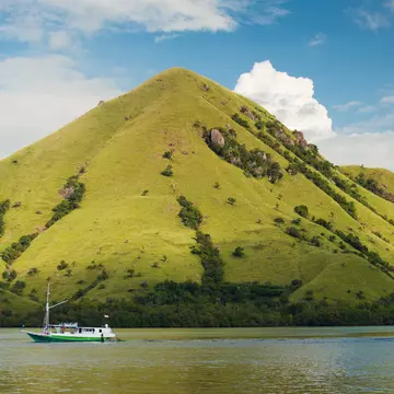 The sun strikes the green grassy slopes of Rinca Island (also known as Rincah and Rindja), as a small boat travels through the water in the foreground.