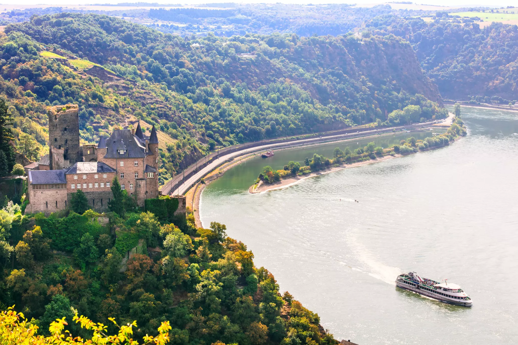 A view of a river from the top of a bluff. A castle is at the top of the bluff in the foreground, while on the water a cruise ship navigates a bend in the river.