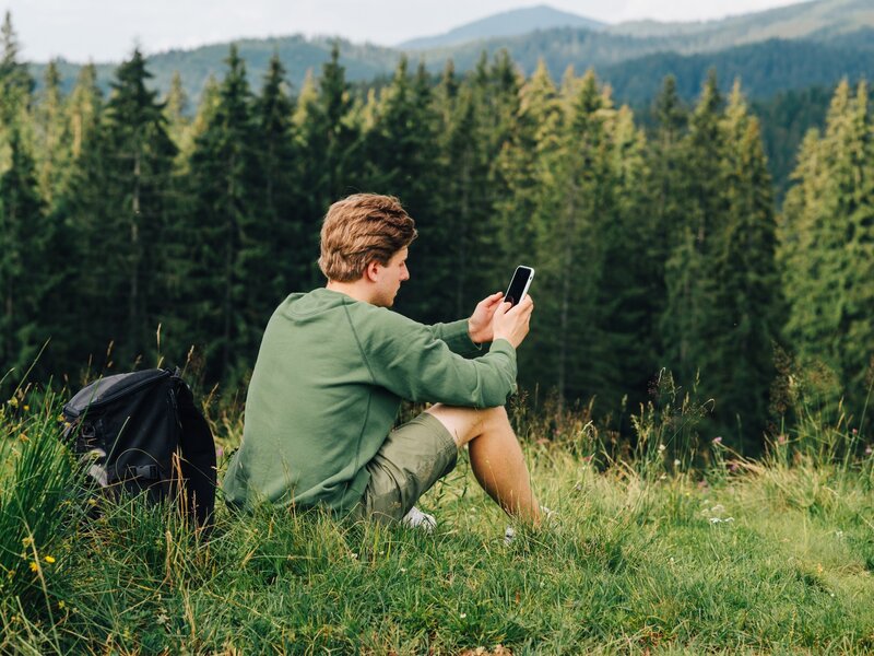Tourist sits on the grass of a mountain meadow with a backpack and uses the Internet on a smartphone with a serious face on a background of beautiful views.