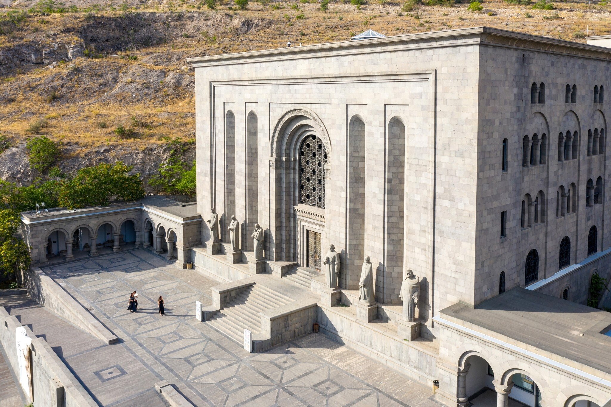 Grey stone building with a tall arched doorway and stone statues along its steps above a paved plaza.