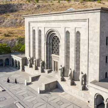 Grey stone building with a tall arched doorway and stone statues along its steps above a paved plaza.