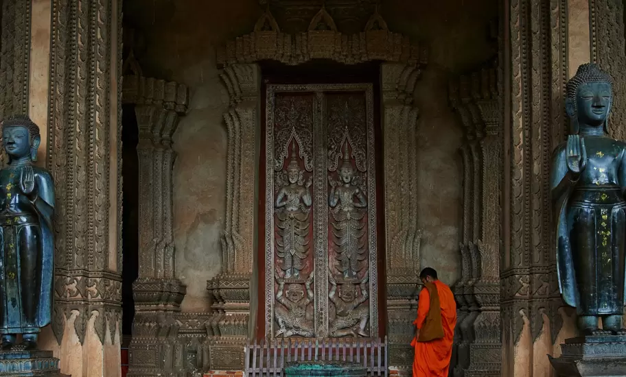 Monk in orange robes at the carved wooden doors of Haw Phra Kaew in Vientiane, flanked by bronze Buddha statues