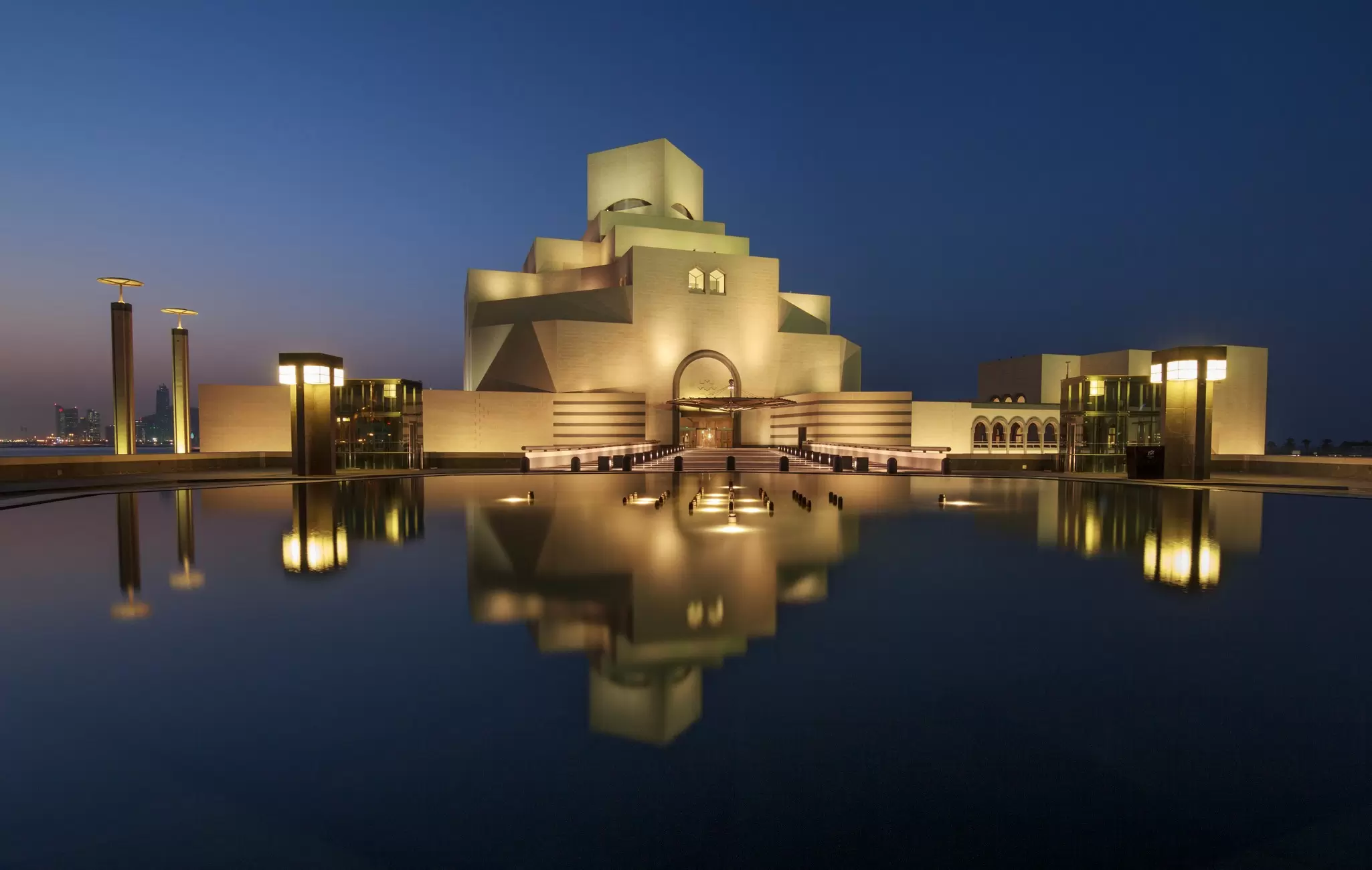 The geometric stone Museum of Islamic Art lit at dusk and mirrored in calm water on the Doha waterfront.