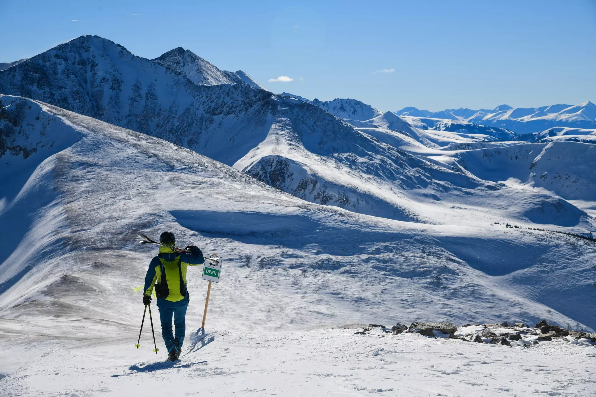 A skier is pictured at the top of a peak, with a view of many snowy peaks in the distance under a bright blue sky.