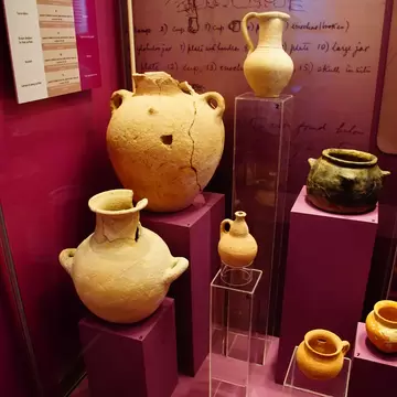 Buff and orange clay jars, jugs and bowls raised on perspex plinths inside a magenta-lined museum case.