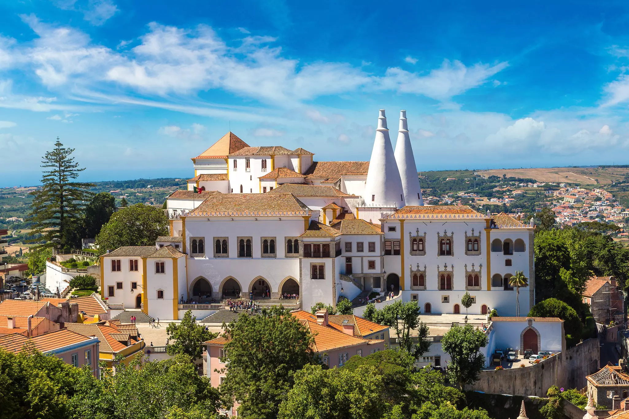 A large palace complex on top of a hill with whitewashed walls, a terracotta-tiled roof, and two conical white turrets.
