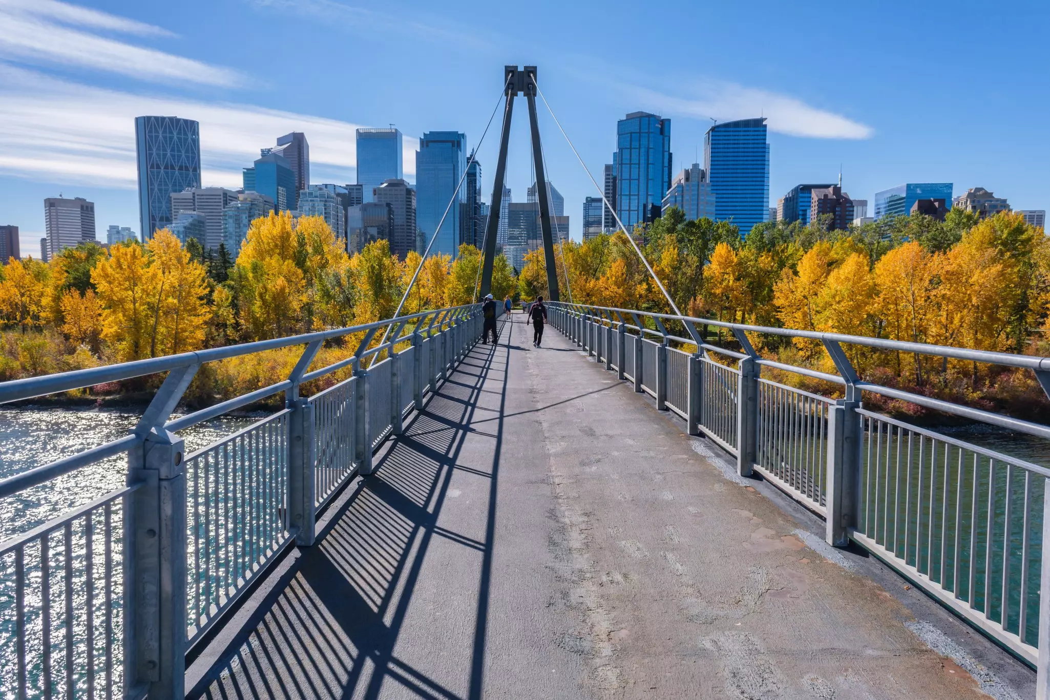 People walk on a pedestrian bridge in a city with towers and trees showing fall foliage.