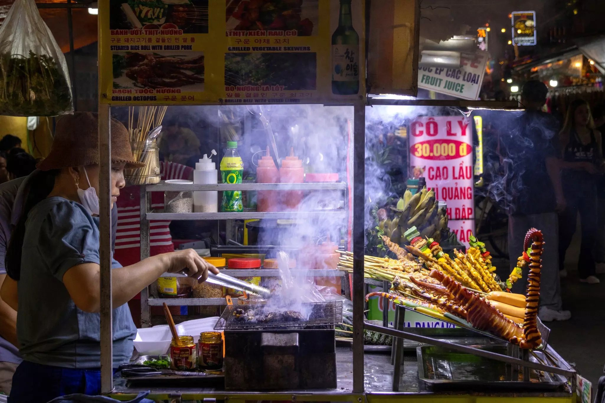 A woman grills meat over a smoky fire at a night market.