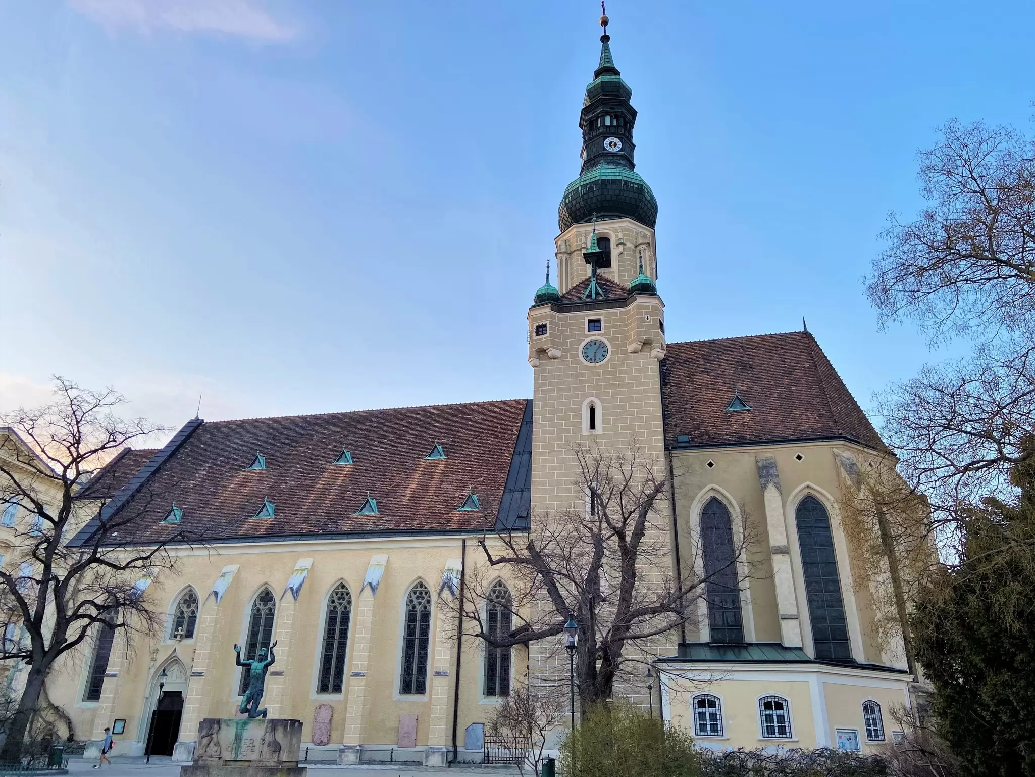 Pale yellow Gothic church with pointed windows and a green copper onion-dome bell tower, framed by bare trees