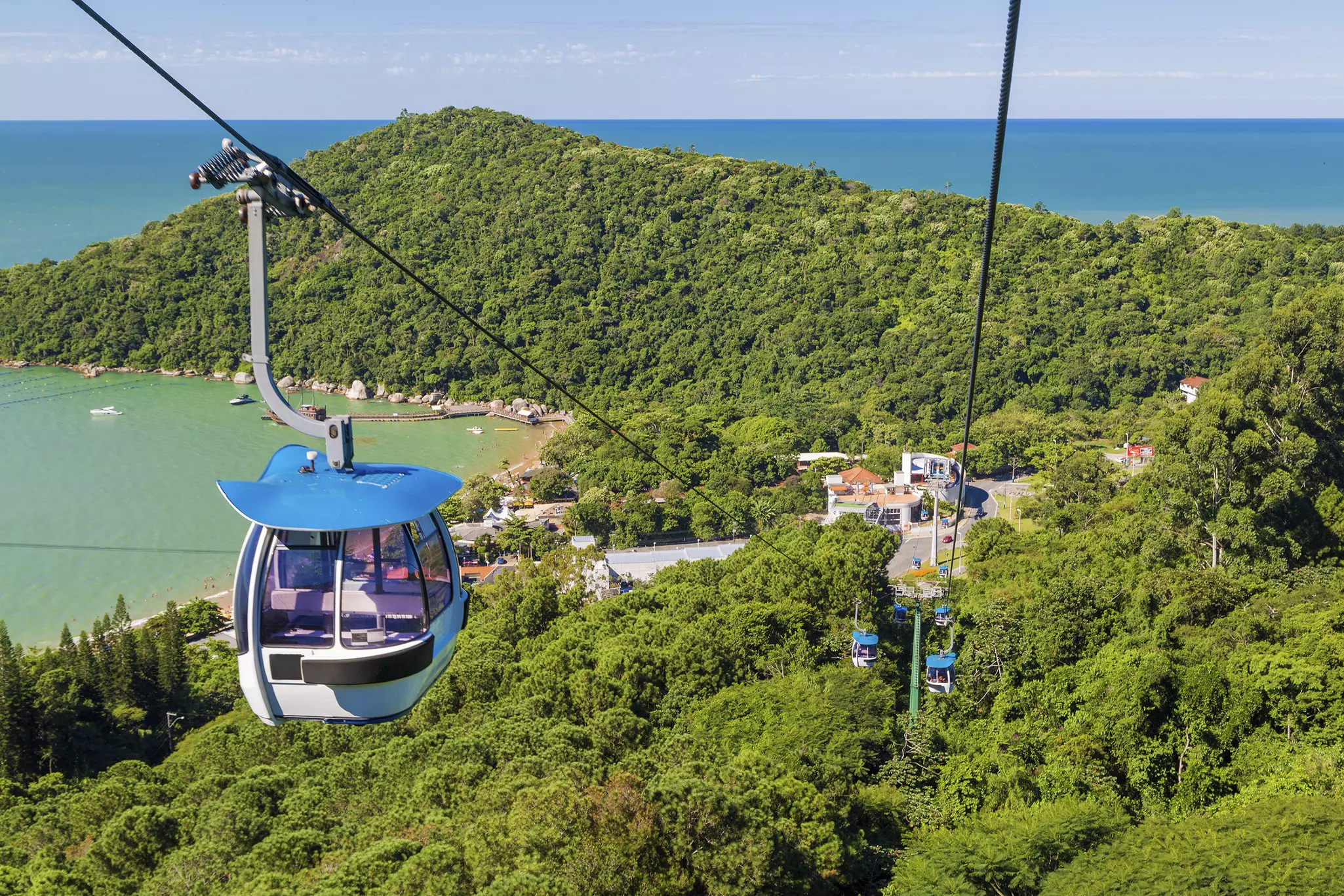 A blue cable car carriage rises above dense woodland in a hilly seaside settlement