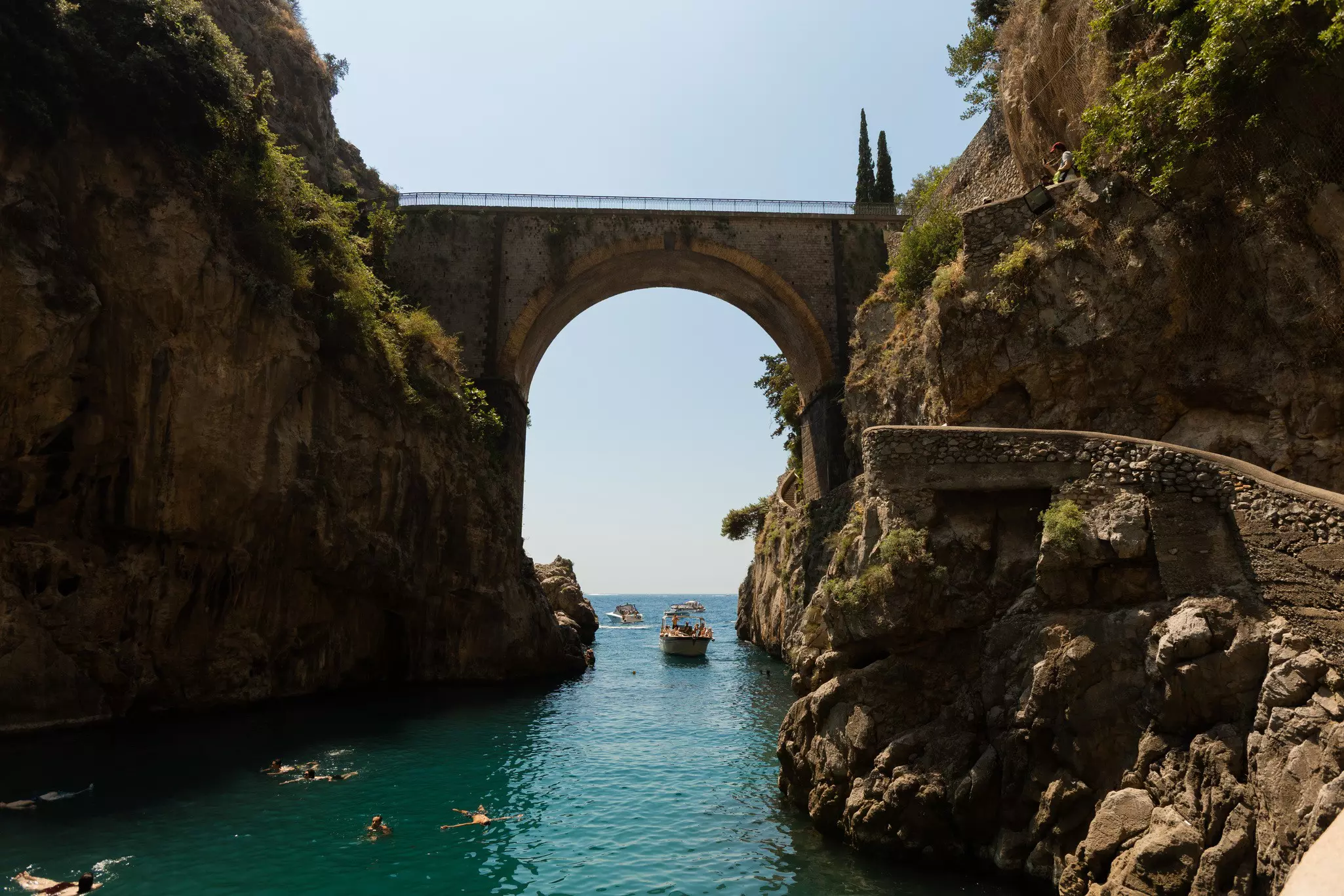 People swim and boat in a cove bordered by rocky hillsides with an arched stone bridge in the distance on a sunny day.