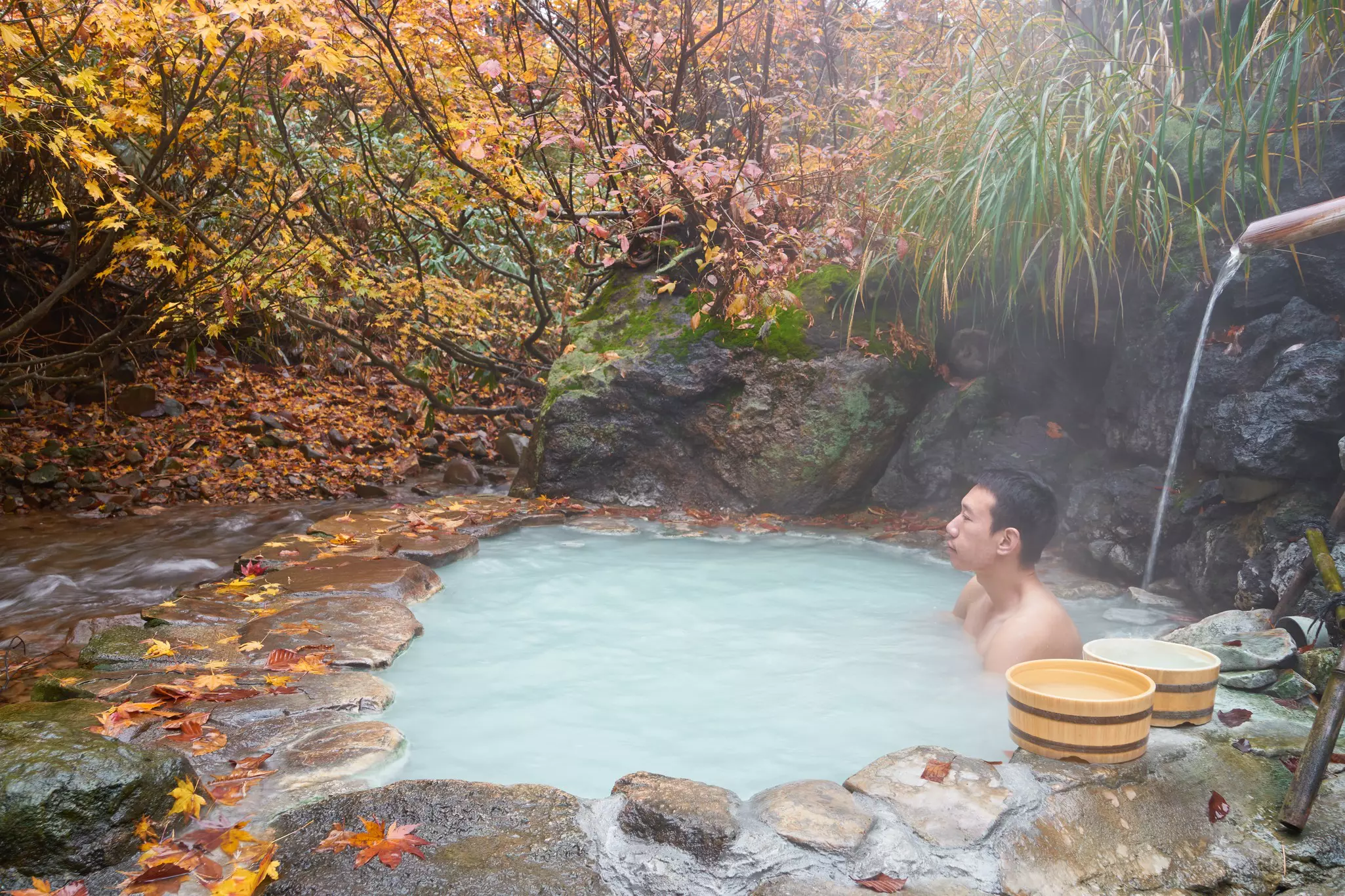 A man soaks in a hot spring under a canopy of fall foliage in a forest.