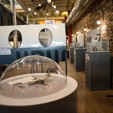 Model aircraft under a clear dome in a stone-walled museum gallery lined with display cases and panels.