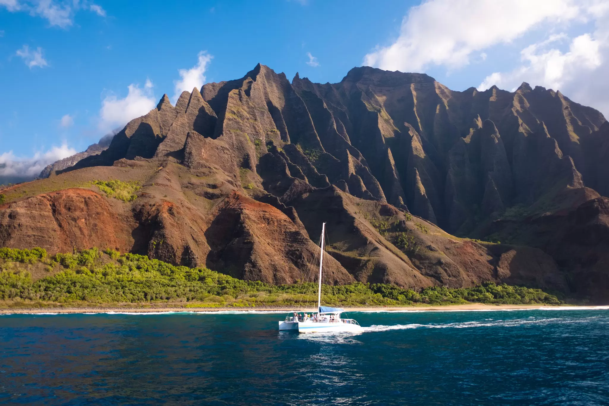 A sailboat in the ocean off the shore of an island with vast pointed cliffs, Nāpali Coast of Kauaʻi, Hawaiʻi, USA