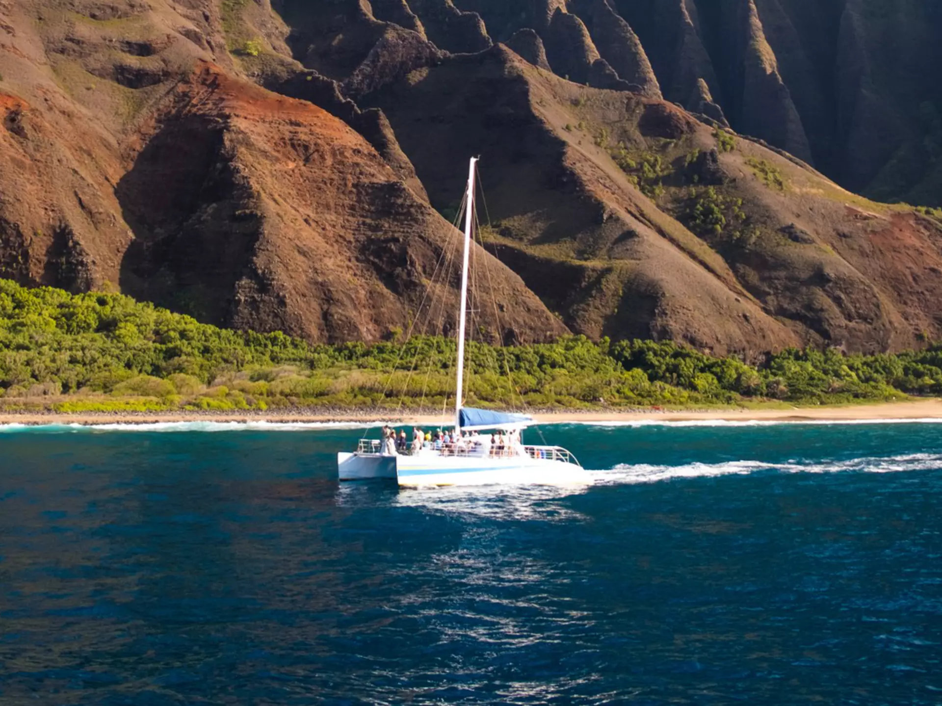 A sailboat in the ocean off the shore of an island with vast pointed cliffs, Nāpali Coast of Kauaʻi, Hawaiʻi, USA
