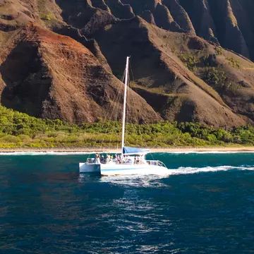 A sailboat in the ocean off the shore of an island with vast pointed cliffs, Nāpali Coast of Kauaʻi, Hawaiʻi, USA