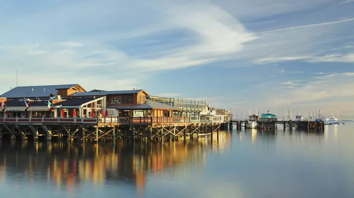 Wharf buildings reflected in the still water of the ocean and lit by an afternoon glow blue sky above dotted with clouds
