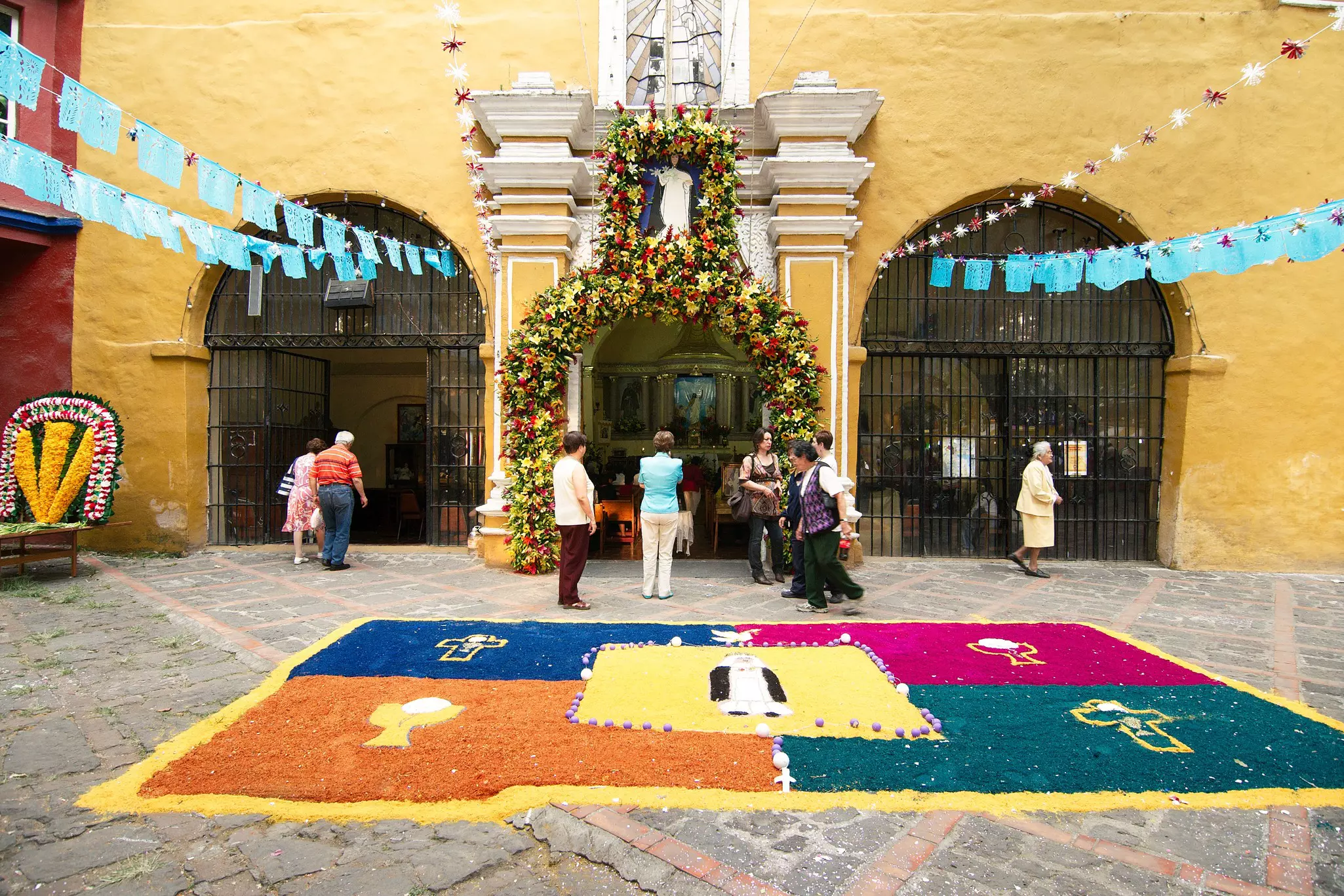 A yellow church has a floral arch around a doorway and a patterned design in the courtyard; blue bunting extends from the church overhead.