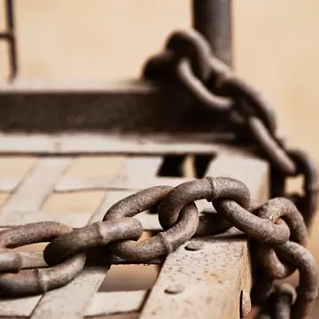 Close-up of thick rusted iron chain links lying across a metal grid bed frame against a plain beige wall