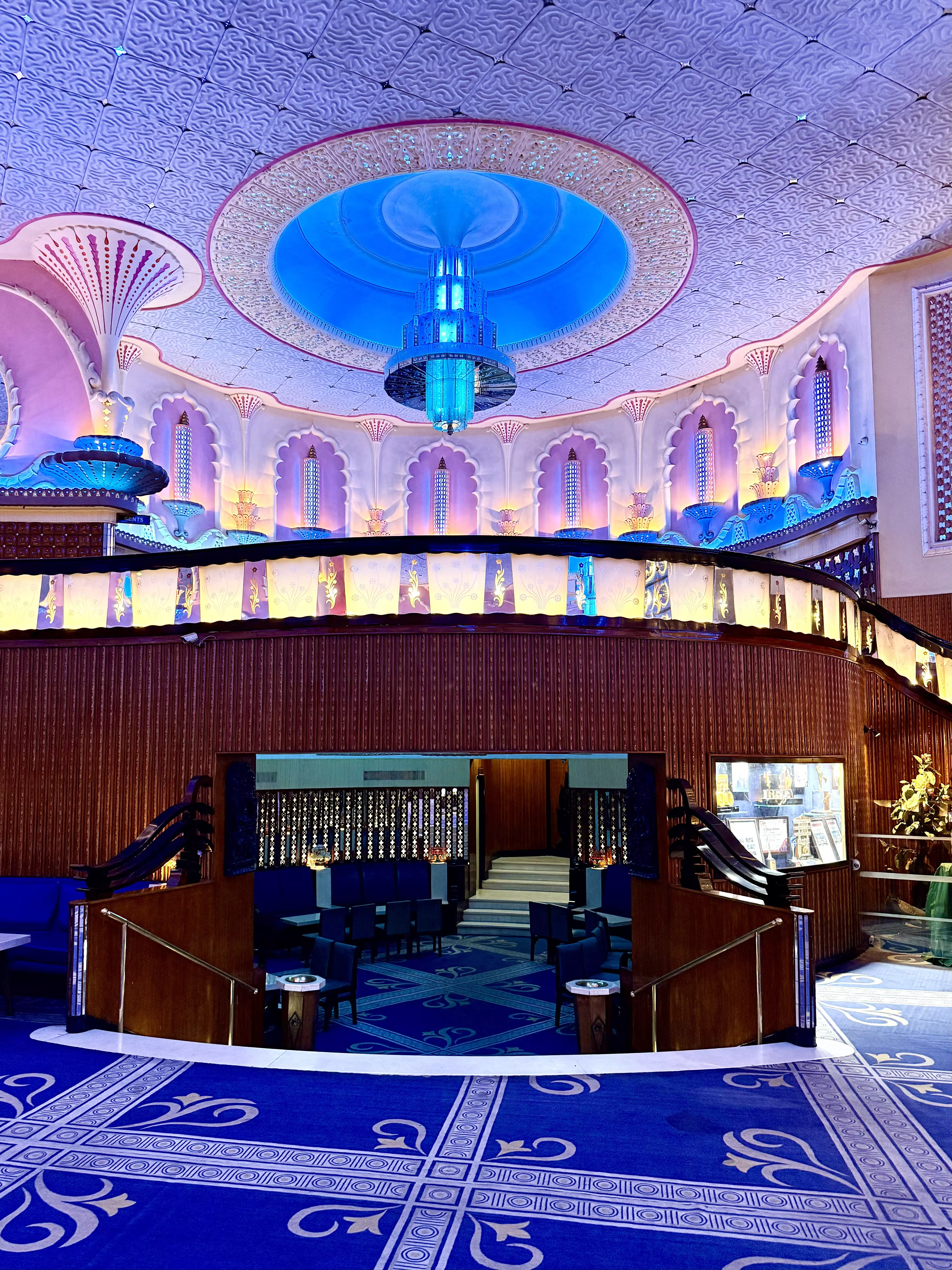 An elaborately designed interior with a blue chandelier and blue light fixtures, walls cast in pale pink light, a blue patterned carpet and dark blue couches in an enclosed space on a lower level.