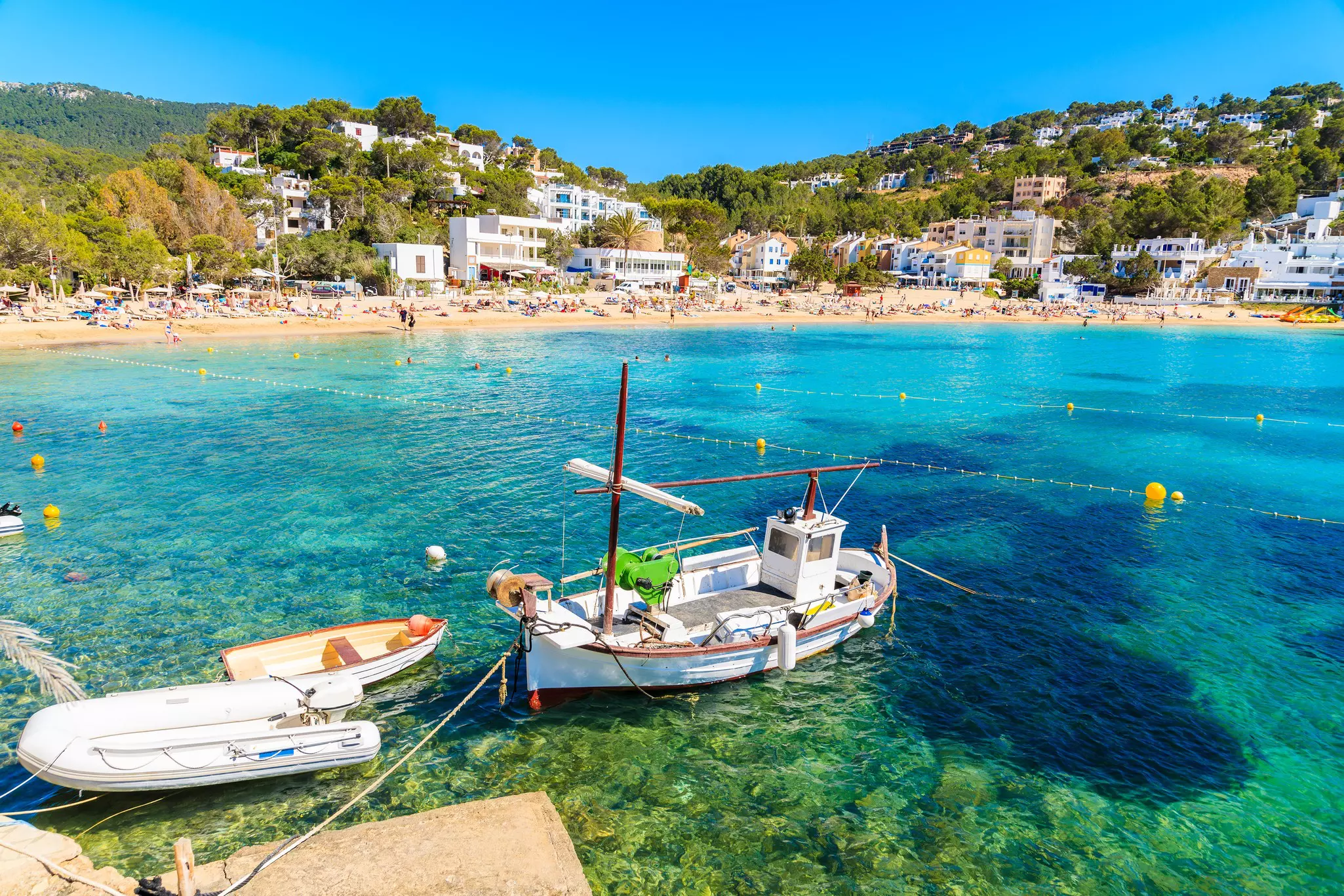 In the foreground, fishing boats are parked in clear water. In the background, rows of whitewashed apartment buildings look out on to a sandy cove lined with sun loungers and beachgoers.
