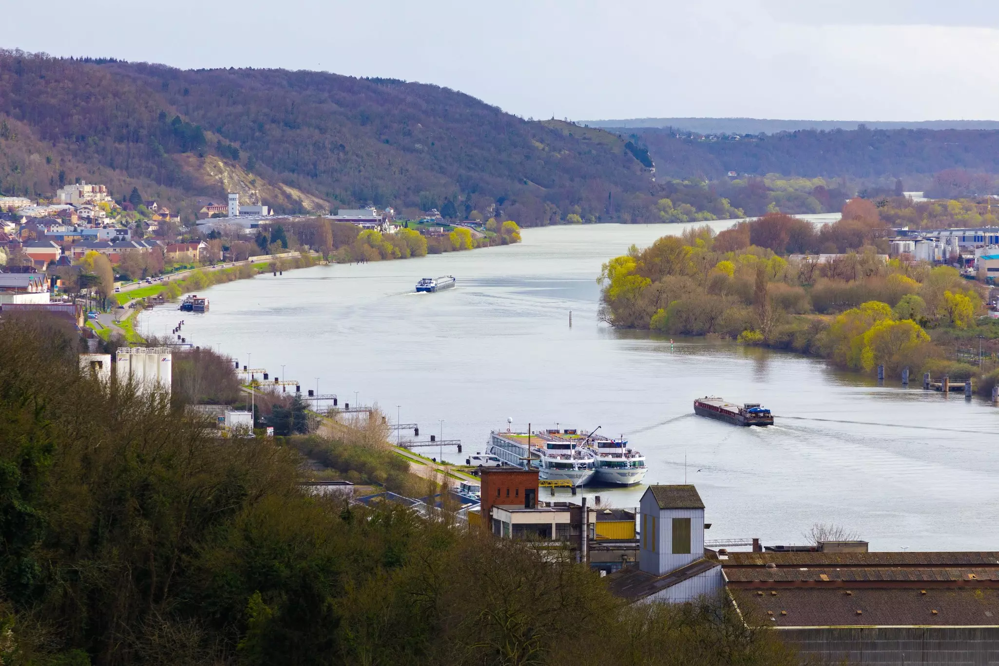 An aerial view of barges and cruise ships on a river in fall. Wooded slopes lead down toward buildings by the riverfront.