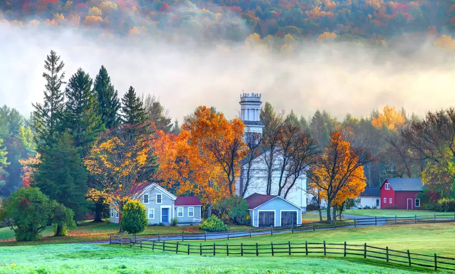White church with a tall cupola amid orange autumn trees and farmhouses in a misty New England valley, eastern USA