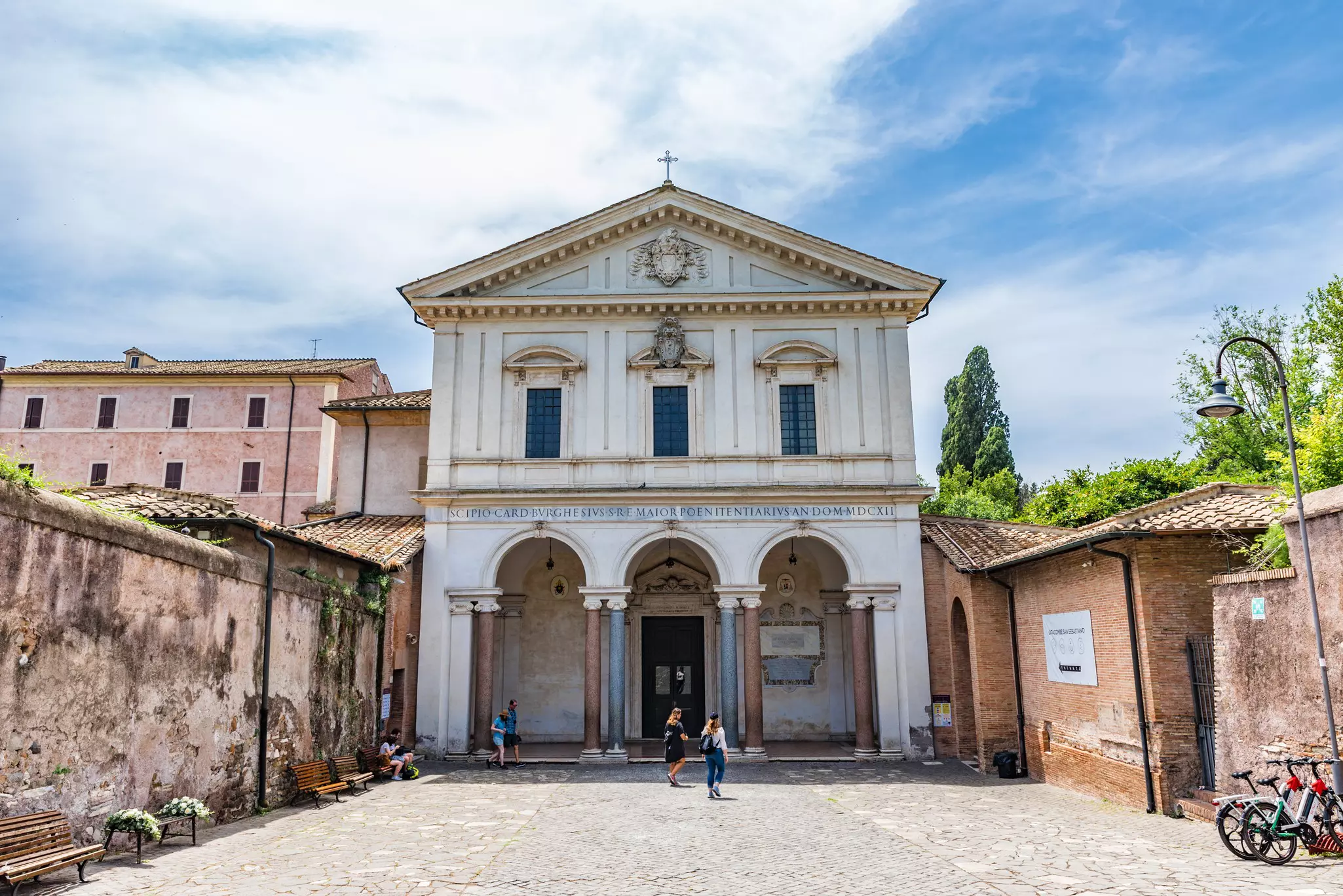 People stand outside the grand entrance to some catacombs, marked by a large white building with columns either side of the door.