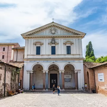 People stand outside the grand entrance to some catacombs, marked by a large white building with columns either side of the door.
