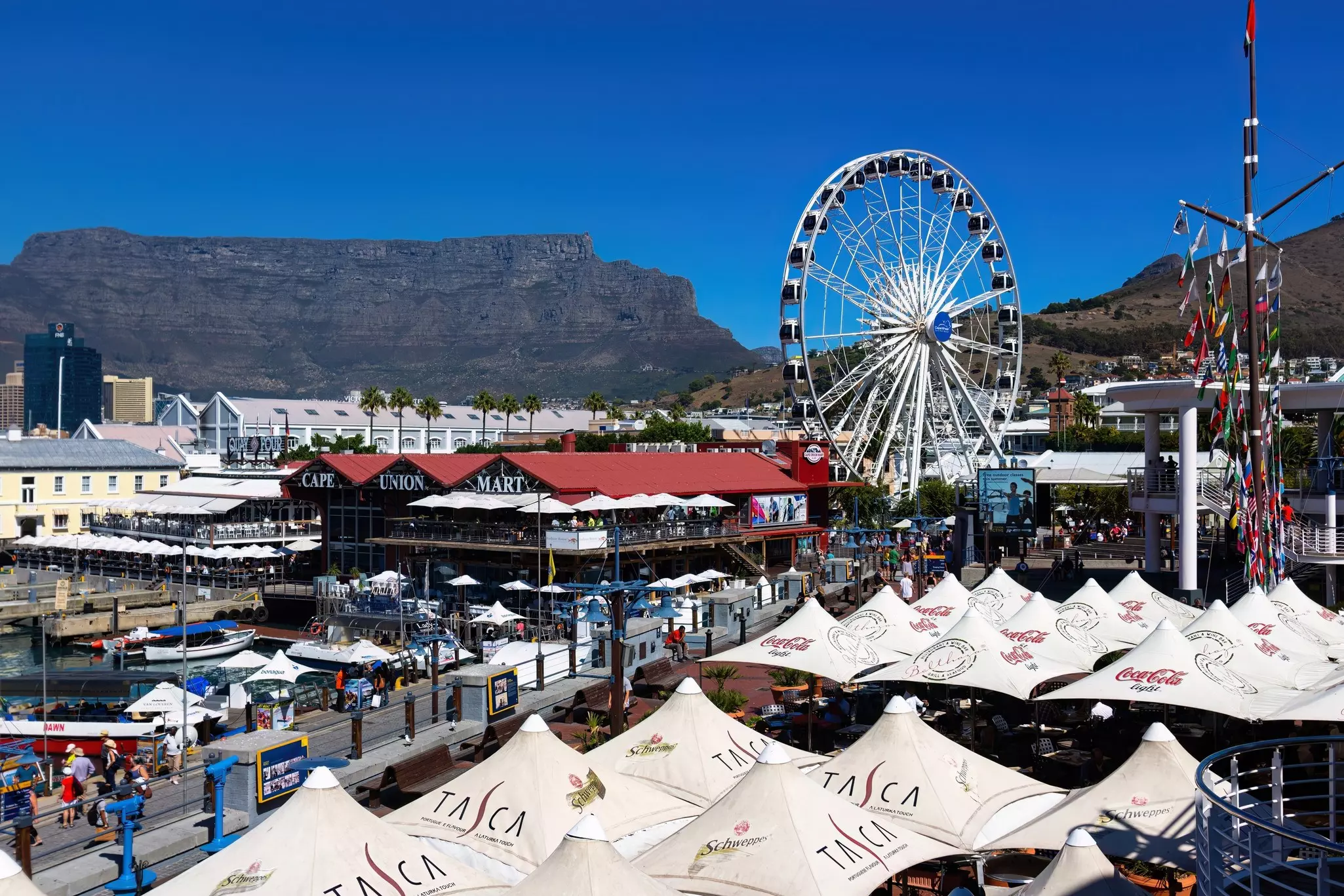 A busy and crowded waterfront with tented booths, a ferris wheel and boats in the harbor