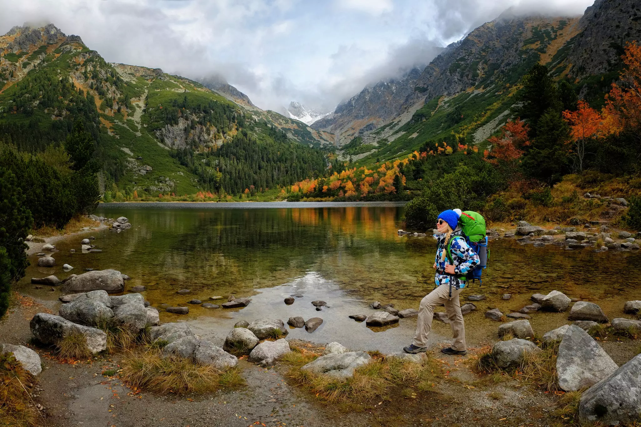 A hiker with a backpack stands by rocks on the edge of a mountain lake in Slovakia, with trees on the shore turning autumn colors.