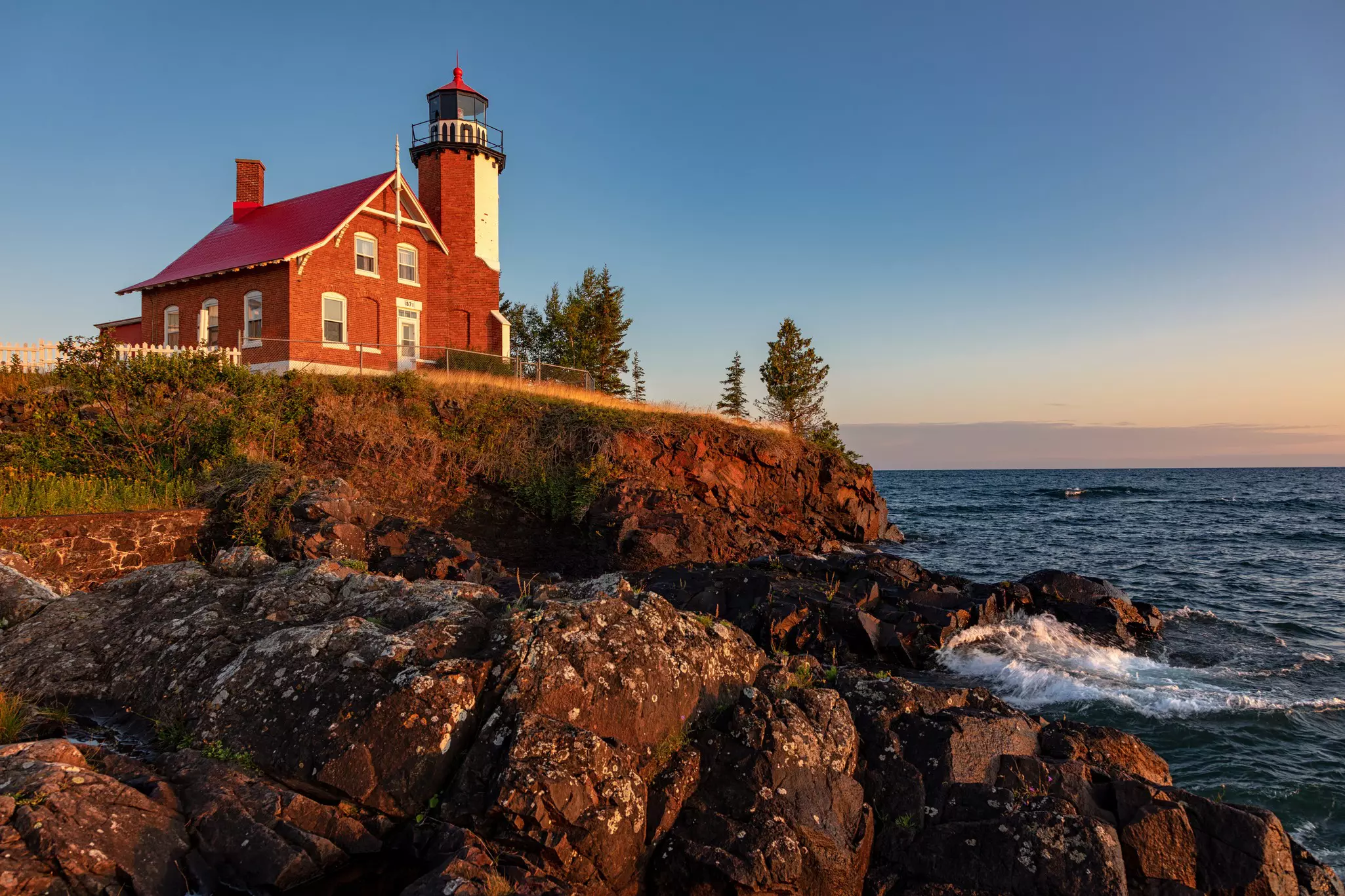 Early morning sunrise illuminates the rocks and front of Eagle Harbor Light on the Keweenaw Peninsula as waves from Lake Superior wash against the rocks.