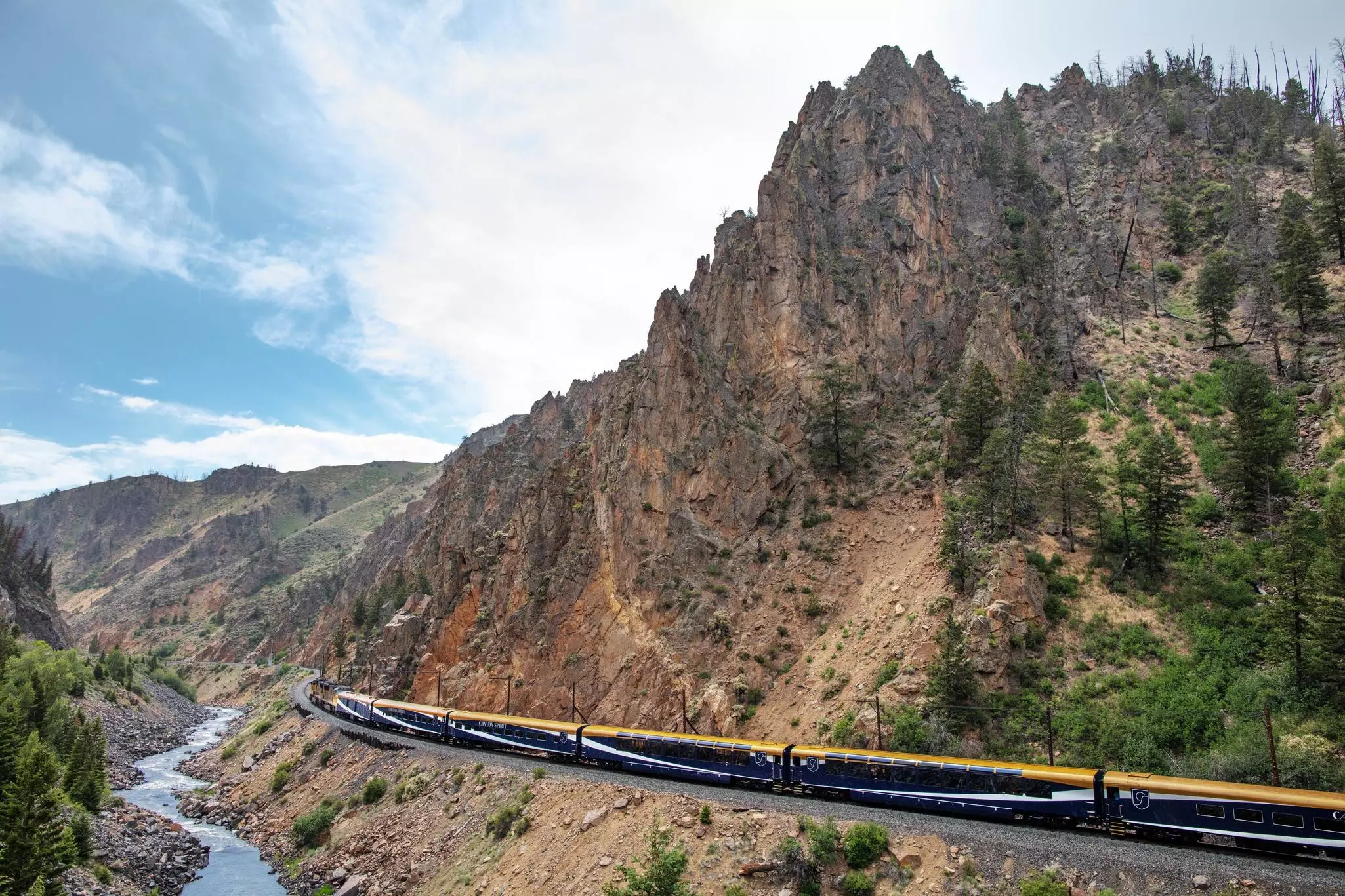 A train on tracks below a low, rocky mountain and a creek on the other side on a mostly sunny day.
