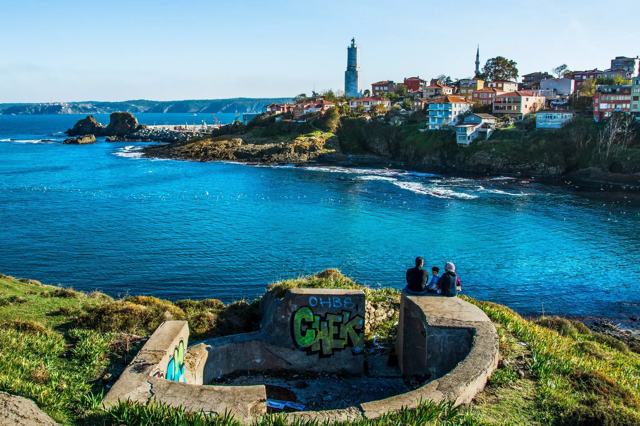 People sit on a defensive concrete structure overlooking a cove, with a lighthouse and village on the far shore.