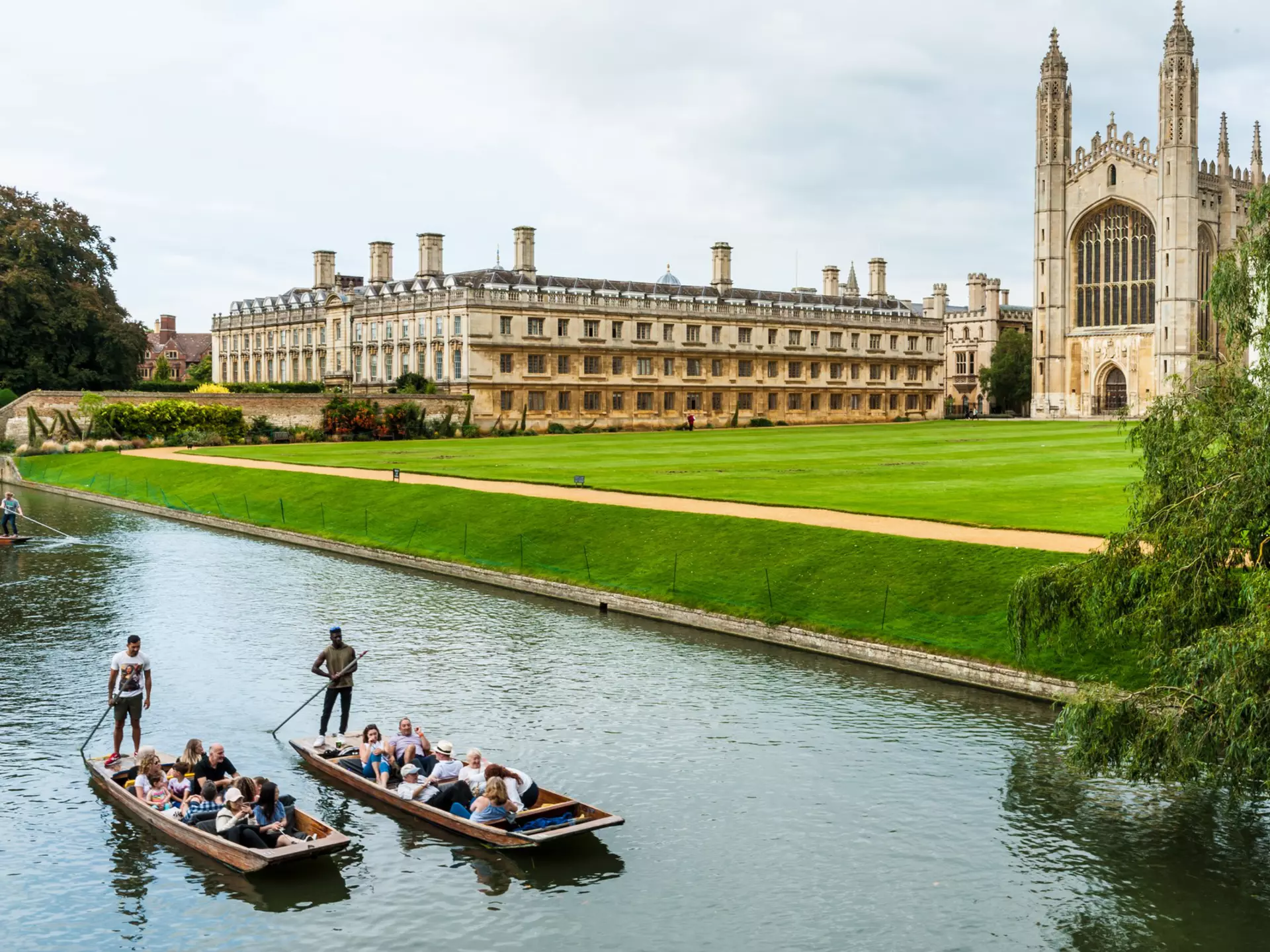 Two small boats are piloted along a river beside a vast green with a large university building and Gothic chapel.