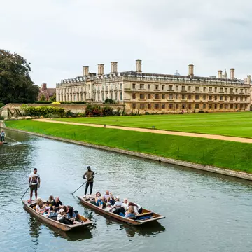Two small boats are piloted along a river beside a vast green with a large university building and Gothic chapel.