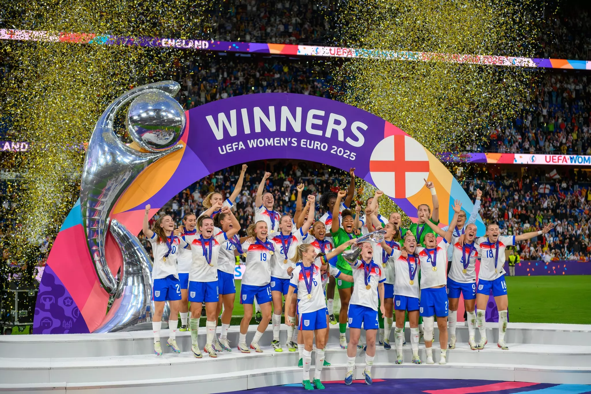 A team of footballers celebrate by lifting their trophy over their heads. They are the winners of the UEFA Women's Euros 2025